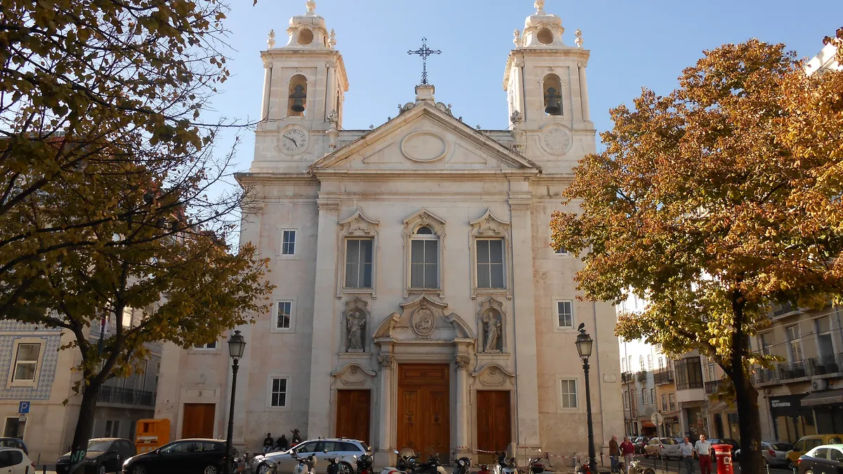 The twin-towered, pale stone front of the Church of São Paulo on its square in Lisbon, with autumn trees either side and tiled houses at the left