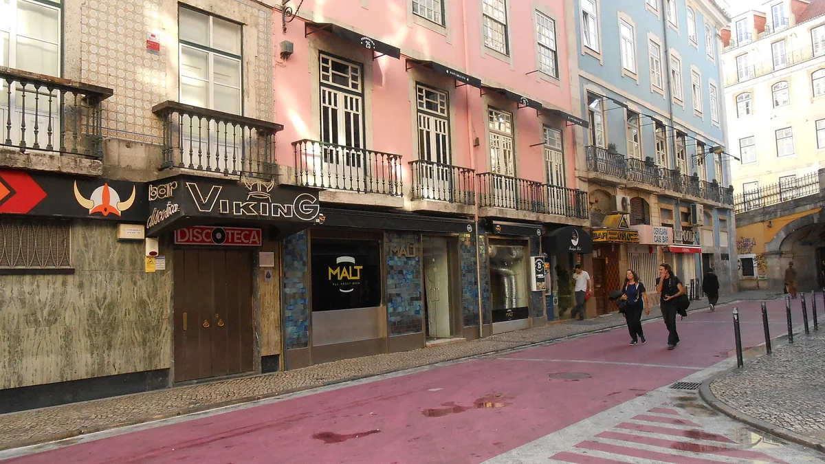 Bars and clubs along a street with pink paving in central Lisbon, shuttered by day, with tiled and painted house fronts above
